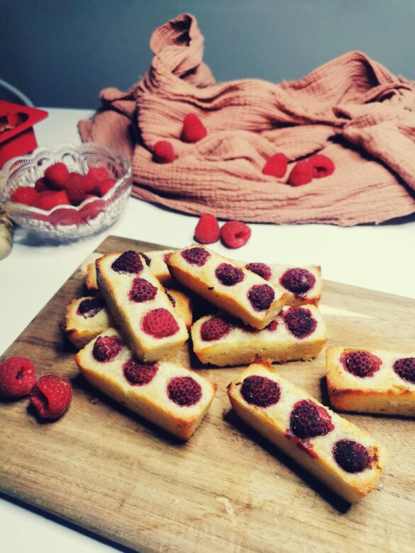 Plusieurs petits financiers à la framboise sur une planche en bois entourés de framboises fraiches dispoées un peu partout